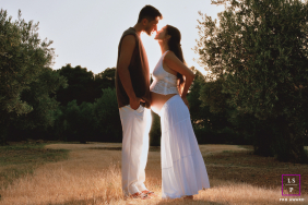 An expectant couple stands face to face in warm backlight, celebrating their pregnancy among olive trees in Jaén, Spain.