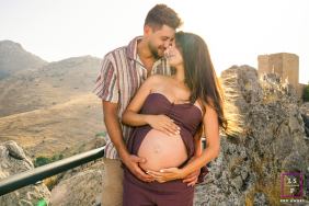 An expectant couple lovingly cradles her pregnant belly while sharing an affectionate connection against the mountain landscape of Jaén, Spain.