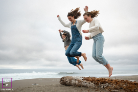 Three Sisters Leap Barefoot From Driftwood Log On Marin Beach Three sisters leap barefoot from a driftwood log together on a beach beneath cloudy summer skies in Marin, California.