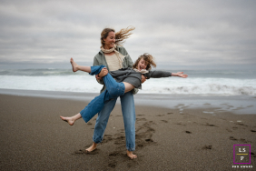 Mother Spins Laughing Daughter Barefoot On Beach In Marin California A mother spins her laughing daughter in her arms while barefoot on a beach beneath cloudy summer skies in Marin, California.
