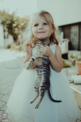 A young girl in a white dress smiles while gently holding a small kitten in her arms outdoors in North Macedonia.