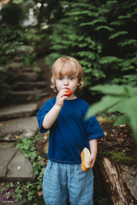 A young boy enjoys a snack while standing among lush greenery and garden pathways during a relaxed family session in Boston.