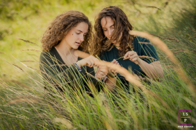 Two Sisters Playfully Explore Tall Grasses Together In Vogelenzang Netherlands Two sisters sit together among tall grasses, playfully exploring nature during a relaxed family session in Vogelenzang, the Netherlands.
