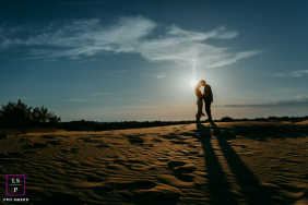 A couple kisses on the sand at sunset, their long shadows stretching across the beach at Plage de l’Espiguette, France.