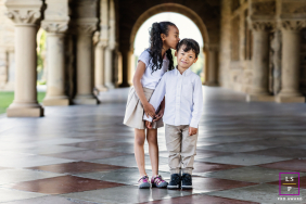 Young Girl Kisses Boy Beneath Elegant Arched Walkway At Stanford A young girl affectionately kisses a boy on the cheek beneath an elegant arched walkway during a family session at Stanford, California.