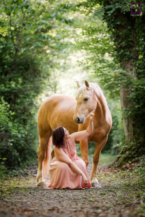 A woman gently reaches toward her horse while sitting together along a lush wooded path in Lyon, France.