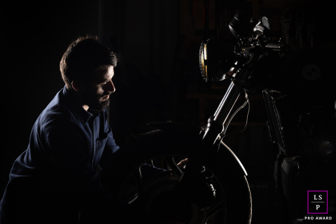 A motorcycle mechanic poses by the front wheel and forks of a bike in Ruleshaker garage, Peyrehorade, France. He’s backlit and rimlit, creating dramatic light around his silhouette.