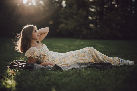 In Champaign, IL, a girl in a floral dress relaxes on the grass, bathed in the golden hour glow. The scene captures a peaceful weekend of sunshine and tranquility, highlighting her serene connection with nature.