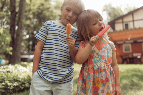 Siblings enjoy popsicles on a warm summer day in the city of Washington. They are laughing and smiling as they sit together, creating a joyful scene.