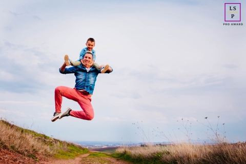Auvergne-Rhone-Alpes dad and son pose for a Lifestyle portrait with the boy on his father's shoulders as dad takes a big leap in the air