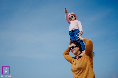 In this picture, a mom lifts her daughter while she points at something far away, against a simple blue sky background in Paris.