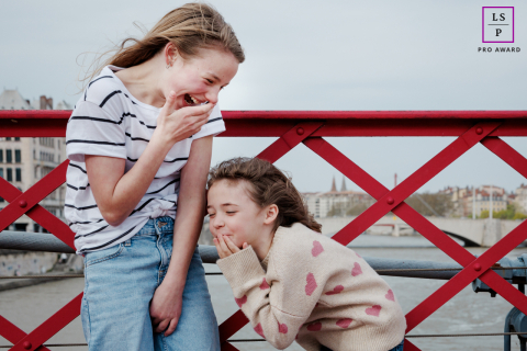 In a windy Paris setting, two sisters share a joyful laugh while standing on a bridge over a canal.