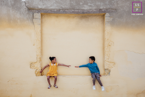 Rennes portrait of two siblings sitting on the edge of a wall, playfully reaching towards each other with big smiles on their faces.