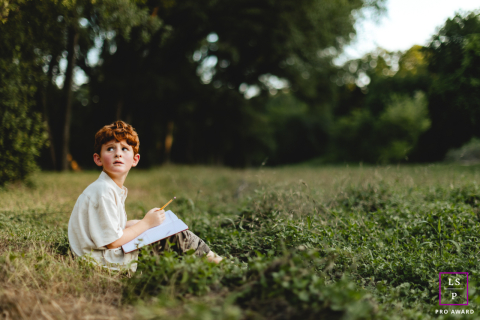 Jeune artiste à Austin : un garçon dessine sur l'herbe verte en regardant en arrière Un jeune garçon d'Austin, au Texas, est assis sur l'herbe verte, absorbé à dessiner dans son livre, son regard doucement tourné derrière lui.