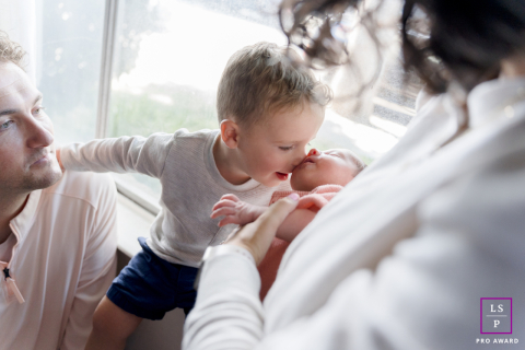 A beautiful photograph of a family, taken in their home on the couch by the window, with the toddler giving his new sibling a sweet kiss.