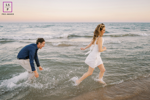A couple playfully splashing each other by Lake Michigan in Sheboygan, Wisconsin. They laugh and run on the water's edge during a sunny day.
