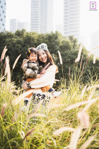 A lifestyle portrait captured in Chengdu, China, featuring a mother and her young toddler. The soft light illuminates their loving bond as they play in tall grass, with the city skyline serving as a backdrop.