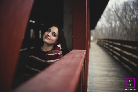 Golden Reflections: Autumn Serenity on Mahomet's Red Bridge A vibrant young woman pauses on a serene autumn day in Mahomet, IL. She leans against a rustic red wooden bridge, adorned with delicate gold necklaces and a striped sweater. Soft light illuminates her calm and content expression.