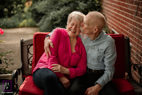 Four Decades of Joy: Celebrating Love on a Mahomet Bench In Mahomet, IL, a couple sitting on a red bench, celebrating their 40th anniversary. The man sweetly kisses his wife's cheek as she laughs with pure joy in their intimate and heartwarming lifestyle portrait. Their bond radiates with love and warmth.