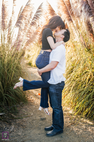 A couple shares a romantic kiss in Santa Cruz, California, surrounded by tall reeds. The man gently lifts the woman off the ground as they share a kiss, showcasing their love and connection. 