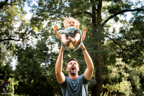 Jeux entre père et enfant dans les bois enchanteurs de la Haute-Garonne En Haute-Garonne, un père soulève joyeusement sa petite fille au-dessus de sa tête, créant une sensation ludique et aérienne au milieu des arbres.