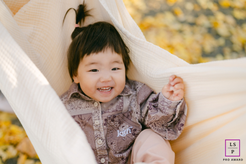 In Hoboken, NJ, a two-year-old toddler girl enjoys the autumn season while playing in a hammock outside. Surrounded by the vibrant colors of fall, her joyful laughter embodies the simple pleasures of childhood.