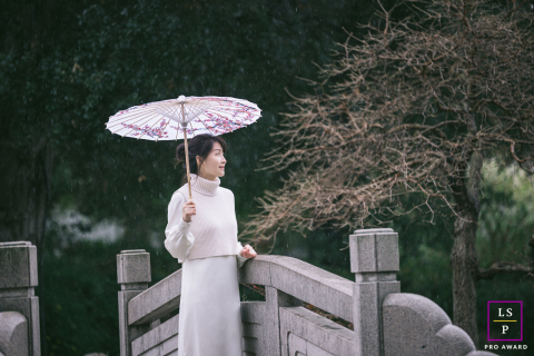 In Los Angeles, at the Chinese Garden in the Huntington Library, a woman holds a traditional Chinese umbrella on a rainy day. The scene captures an incredibly poetic and serene moment amidst the lush garden surroundings.