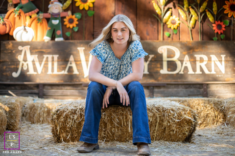 In Avila Bay, CA, a senior student sits on a bale of hay by a barn on a windy day. The rustic setting and breezy atmosphere captures contemplation and connection to the countryside.