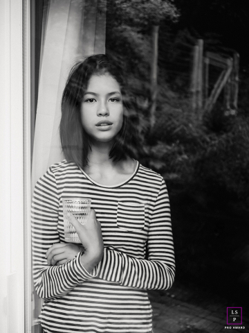 Morning Reflections: A Girl's Quiet Contemplation in Braeside's Soft Summer Light In Braeside, Ontario, Canada, a girl is captured in a black and white shot during an early summer morning, standing in front of a window. The image reflects quiet reflection and soft natural light.