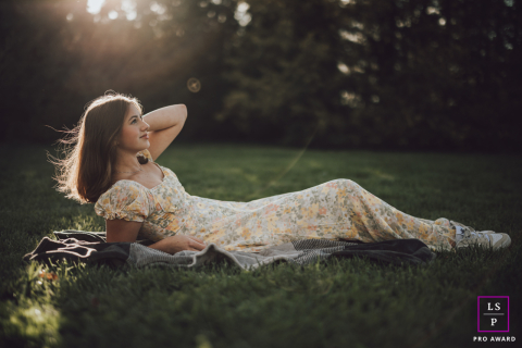 Golden Hour Serenity: Girl's Tranquil Connection with Nature in Champaign's Floral Bliss In Champaign, IL, a girl in a floral dress relaxes on the grass, bathed in the golden hour glow. The scene captures a peaceful weekend of sunshine and tranquility, highlighting her serene connection with nature.