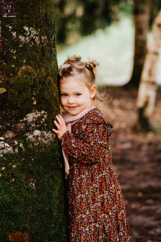 A shy girl is peeking from behind a large tree trunk at Lac de Montriond. The portrait captures her innocent and playful curiosity as she looks out.
