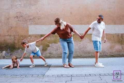 In Bayonne, France, a family walks in single file along the sidewalk: mom, dad, and boy holding hands. The boy reaches back to connect with a seated toddler, emphasizing togetherness and care.