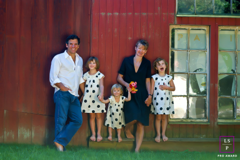 Hamptons Polka Dot Dreams: Um retrato lúdico de família por Red Barn Nos Hamptons, Nova York, uma família posa alegremente perto de um prédio rústico vermelho. A mãe e o pai estão de pé enquanto suas três filhas, todas em vestidos de bolinhas combinando, adicionam charme e energia à cena.