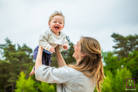 Felicidade pura: a ascensão alegre de uma criança de Hilversum acima da linha das árvores Em Hilversum, na Holanda, uma criança cai na gargalhada enquanto sua mãe a ergue bem acima da linha das árvores. Sua expressão alegre e a vista deslumbrante transmitem uma sensação de liberdade e deleite.