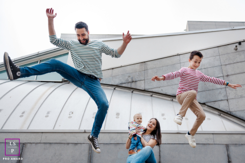 À Paris, le saut joyeux d'une famille vers une vie audacieuse et graphique À Paris, une famille joue ensemble : un père et son fils sautent à l'unisson, filmés en plein vol. En arrière-plan, une mère et un autre enfant jouent près de bâtiments aux formes graphiques et audacieuses, ajoutant une énergie dynamique à la scène.