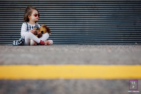 Le calme d'un enfant dans une rue industrielle de Metz À Metz, une petite fille est assise sur le béton près d'une porte métallique industrielle, câlinant son jouet préféré. Des lignes de signalisation jaunes marquent le premier plan, et la contre-plongée souligne sa petite présence pensive.