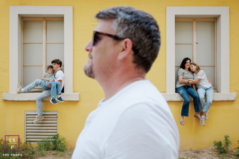 Un portrait de famille parisien élégant : papa de profil, famille en couches À Paris, un portrait de famille présente le père au premier plan, de profil, tandis que le reste de la famille est assis sur le rebord de la fenêtre derrière lui. La composition met en valeur les liens familiaux grâce à un agencement élégant et complexe.