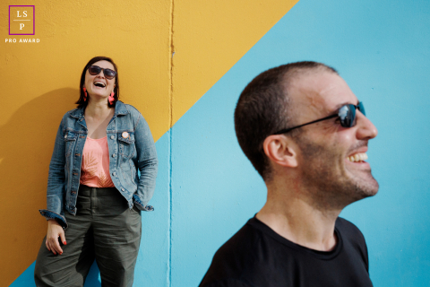 Un portrait graphique de Paris capture l'étreinte joyeuse et radieuse d'un couple À Paris, un couple pose pour un portrait graphique. Il se tient au premier plan, de profil, tandis qu'elle est derrière lui, dos au mur. Tous deux arborent un large sourire, rayonnant de joie.