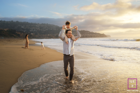 At Redondo Beach, CA, a mother and father play with their two sons on the beach at sunset. The family is surrounded by joyful laughter, their smiles glowing softly in the golden light.