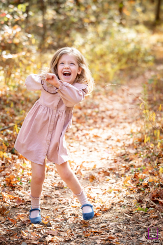 In Alexandria, Virginia, a little girl stands among fallen leaves, making a playful dancing gesture with a big, joyful smile, delightfully capturing the fun and energy of autumn.