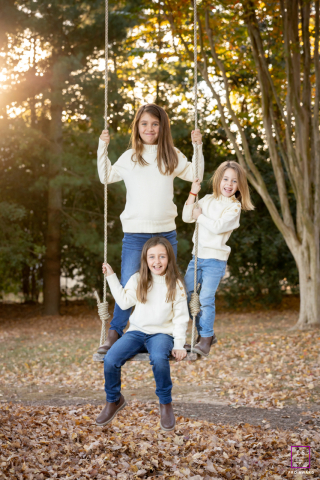 In St. Michaels, Maryland, three girls smile and pose while sitting together on a rope and wood plank swing hanging from a large tree, all looking warmly at the camera.