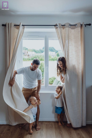 In Solaize, Rhône-Alpes, France, a family plays hide-and-seek behind window curtains. Playful laughter is revealed through the light filtering softly around them, capturing a joyful indoor scene.