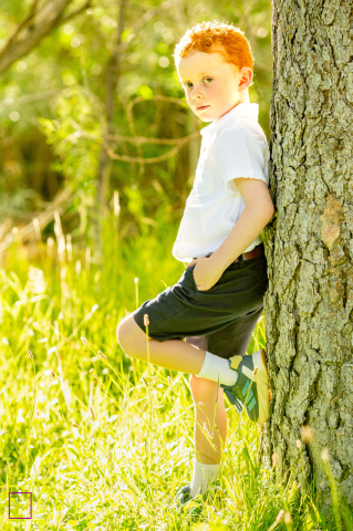 In Reno, NV, a boy poses stylishly, leaning against a tree in a grassy field during golden hour. Backlit by warm sunshine, the scene glows with soft, golden tones and highlights his playful attitude.