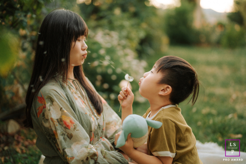 At Shinn Historical Park and Arboretum, a child blows dandelion seeds toward their mom’s face under the warm glow of sunset, creating a whimsical and tender outdoor scene.