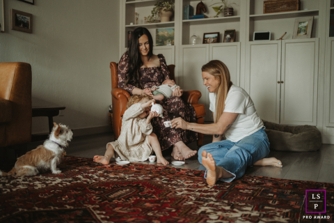 In Portland, OR, two women sit in a cozy living room enjoying tea time with young kids and a dog, all gathered together and sharing a warm, relaxed scene.