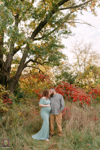 Waukesha Autumn Embrace: Parents-to-Be Share Tender Kiss Amidst Vibrant Foliage In Waukesha, Wisconsin, parents-to-be share a quick kiss among vibrant fall foliage, captured in a vertical shot surrounded by autumnal nature.