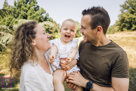 In Lyon, France, mom and dad carry their daughter outdoors, laughing together and surrounded by nature.
