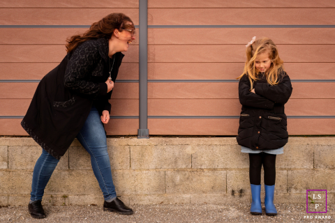 In Annecy, Haute-Savoie, France, a mother and daughter laugh together, playing with their hair on a windy, chilly day in an urban setting.