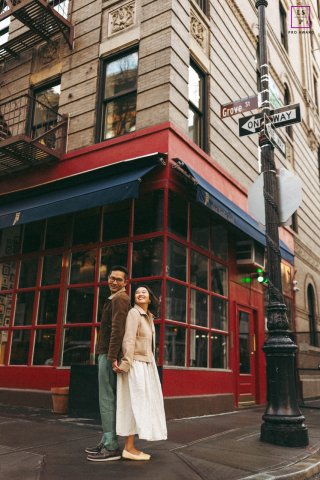 Outside Little Owl in West Village, Manhattan, a couple stands back to back, both smiling, framed by the lively city street.