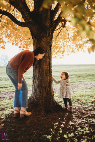 Mother and daughter playing peekaboo near a tree in Boston, capturing a tender lifestyle moment.